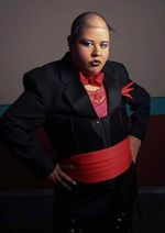 Koomah with an intense expression and their arms akimbo. Koomah is an Asian/Latinx person with a buzz cut, light skin and dramatic makeup. They wear a blazer, corset, cumberbund, lace top, and bowtie.