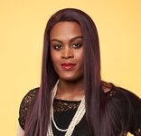 Mya Taylor is a Black trans woman with dark brown skin in front of a yellow background. She is wearing red lipstick and has long dark red brown hair. Mya is wearing a black lace short sleeve top and a white pearl necklace.