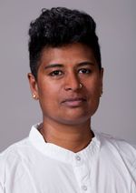 Neelu Bhuman is a nonbinary trans masc South Indian person with dark brown skin wearing a white button up. They are in front of a light grey background. They have gold earrings and a nose ring. They have short black hair with it faded on the sides.