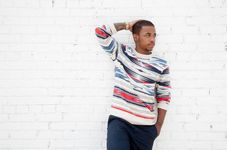 Seven King is a Black trans man in front of a white brick wall with brown skin. He has a multicolored crew neck sweatshirt with navy blue slacks. He has one hand in his right pocket and the other hand resting on the back of his head.