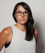 Sydney Freeland is a Navajo trans woman with light brown skin in front of a white background. She is wearing a white tank top with a black undershirt peeking out from underneath. She has long black hair pulled onto her left side flowing onto her chest. She is wearing black eyeliner and black glasses.