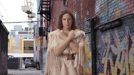 Film still from Woman Dress. An Indigenous person with long wavy brown hair, wearing a light-brown fringed leather dress lined with fur and a beaded belt stands looking at the camera with their arms forming an X in front of them, their hands closed into fists. They are standing in an urban alleyway beside a colorful graffitied brick wall.