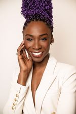 Angelica Ross is a Black trans woman with dark brown skin. She is posing in front of a white background wearing a white blazer. She has her hair in purple box braids and they are fashioned into a bun. She is resting her right hand on her cheek.