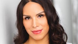 Kiley May is a Two-Spirit transgender person with light brown skin. She has curly brown hair and is posed in front of a blurred gray background.