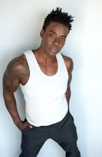 Skyler Cooper is a Black trans man with dark brown skin. He is posed in front of a white background in a white muscle t-shirt with his hands in their pockets. He is wearing black dress slacks and has black hair with the sides short.