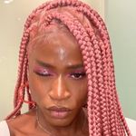 Miles Jai is a Black nonbinary trans femme person with dark brown skin posed in front of an off white background. She has pink box braids with pink eyeshadow and a white t-shirt.