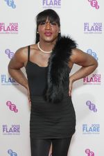 Kristen Lovell is a Black trans woman posing with her hands on her hips in front of a white banner. She has long straight black hair tucked behind her ears with bangs. She is wearing a pearl necklace and earrings and a black dress with black tights and black fur over her left shoulder.