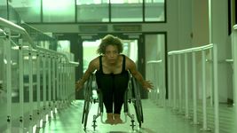 A person with light brown skin, hazel eyes, short light brown curly hair is in a gray wheelchair. They are wearing a black tank top and black leggings with their feet exposed. They are leaning forward, chest to their knees, with a determined expression and their hands on either side of the chair behind them, suggesting that they are in motion. They are in a building with white railings on either side.