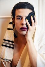 A person looks into the camera shirtless with short black hair and dark purple red lipstick. They are holding a yamaka over their left eye and have yellow nail polish. They have a white and black tallit resting on the back of their head and going down on their shoulders.