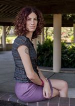 A person is sitting on a short concrete wall. They are looking over their right shoulder with their legs crossed and their hands resting in their lap. They are wearing a lavender pencil skirt and a short-sleeved gray-striped blouse. It is striped with a navy blue and gray pattern.They have shoulder-length curly brown hair with dark brown roots. On their right arm, they have a tattoo with some kind of circular pattern up near their shoulder, and they wear a pink plastic hair elastic near the wrist.