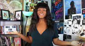 Kat Blaque, a Black person with medium skin and dark wavy hair with blunt bangs, stands in front of a heavily-postered wall with her arms raised in a questioning shrug.