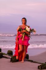 Tourmaline, a brown-skinned person with dark, wet, shoulder-length hair, stands in front of a sunset in a pink ruffled dress, her left foot on the beach and right foot slightly lifted. She holds a bouquet of tulips in her left hand, holding up her dress with her right hand.