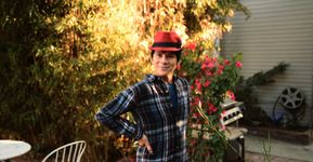 Hida Viloria is a Latine person with brown eyes, short dark hair and light skin, wearing a plaid shirt and a red fedora. They stand on a leafy patio with their hands on their hips and a slight smile on their lips.