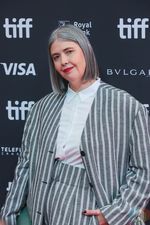 Chelsea McMullan, a white nonbinary person, giving a closed-mouth smile on a red carpet at the 2023 Toronto International Film Festival. They are wearing a grey-and-white-striped oversized pantsuit that compliments their silver shoulder length hair and bright red lipstick.