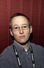 Jenni Olson, a white gender nonconforming butch person, stands in front of velvet red curtains for a photograph, likely for media coverage.