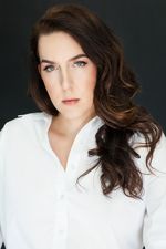 Professional studio portrait of Luna Ferguson, who has light skin, long brown hair and green eyes. They wear a white button-up shirt and stand in front of a dark studio wallpaper.