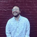 Jamie DiNicola is photographed candidly during a moment of joyful laughter, standing in front of a red brick wall. They have olive skin, short, dark hair and a beard, and are wearing a light-colored button-up shirt and round glasses.