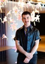 Joseph Liatela has short dark hair and a mustache, and is wearing a black leather buttoned shirt and a print scarf. He is standing in front of an art installation of hanging pink lilies with a calm and confident expression.