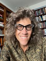 Susan Stryker smiles in a selfie portrait while sitting in a cozy room lined with dense bookshelves. She has shoulder-length curly hair and is wearing distinctive cat-eye glasses with a patterned frame paired with a dark gold-patterned shirt.