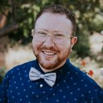 Ewan Duarte smiles warmly at the camera. He has fair skin, short reddish-brown hair and beard, and is wearing glasses, a blue polka-dotted shirt, and a light-colored bow tie. The background is an outdoor setting with trees and greenery, creating a pleasant and natural atmosphere.