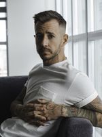 Portrait of Chase Joynt seated on a couch wearing a white t-shirt with his tattooed arms and hands resting across his lap. He has short, well-groomed brown hair and light facial hair. Behind him is a bright window, filling the room with natural light and casting gentle shadows on his face.