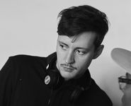 Black-and-white photo of Rhys Ernst gazing contemplatively off to the bottom left. He has short dark hair and a small mustache. He wears a dark long sleeve jacket with black headphones resting around his neck, which provides an aesthetic contrast to the plain light wall and light musical equipment. In the bottom right corner of the image, a sliver of another person's face can be seen. This person has dark skin, glasses, and wears a baseball cap which juts out a little farther into frame.