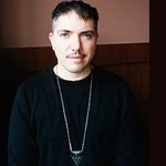 James Diamond stares into the camera in front of a beige-and-brown wall. He has fair skin, short dark hair and a mustache. He is wearing a black long sleeve shirt and long pendant necklace.