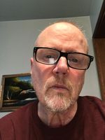 Selfie of Raymond Rea taken in his home looking downwards at the phone camera. He has fair skin, a short graying beard and balding hair, and is wearing a maroon shirt. Behind them is a plain wall with a framed painting.