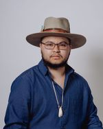 Image of Raven Two Feathers, a masculine Native person in casual western wear and a buffalo tooth necklace, standing in front of a plain white background and staring deeply at the camera.