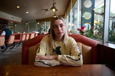 Portrait of Ava Benjamin Shorr in a classic American diner setting. Ava has fair skin, blonde hair and brown eyebrows. Ava is wearing a yellow long sleeve graphic shirt with gold jewelry, complimenting the warm bronze and coral tones in the diner seating and lighting. The lighting is very natural coming from large windows on the right side, leaving Ava's face partly in shadows from the overcast daytime light.