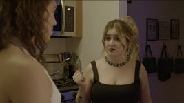 Love is confessing her attraction for Jay to Alex. The two stand in a modern kitchen. Love has curly brown hair and light brown skin, and is seen from over the shoulder. Alex has dirty blonde hair in a ponytail, light skin, long black nails, faces towards camera, and wears a black dress with a necklace of green emeralds.