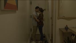 Rickey and Love embracing each other. The are seen from a distance, standing in front of the door of an apartment. They are wearing casual clothes with their feet bare; their shoes are strewn about on the floor beside them.