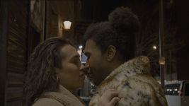 Love and Jay embracing each other in closeup profile, their eyes closed. They are standing on a city sidewalk at night, both wearing fur coats.