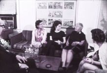 Washed out, grainy 16mm film still. We see three women sitting on a couch, another woman sitting on a chair leaning toward them on the right, and a man with dark glasses sitting on a chair facing them on the left.