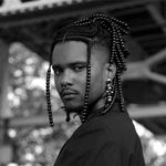 Chester looks over their shoulder while standing under a bridge. They are a Black person with shoulder length braids with metalic beads, a short mustache, and a septum piercing. They wear a black button-up shirt and a dangling earring.