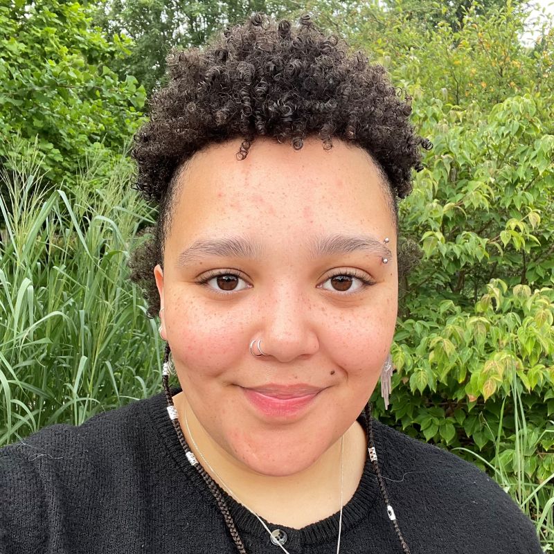 Andi has brown skin and short curly hair. They are pictured with a curt smile.