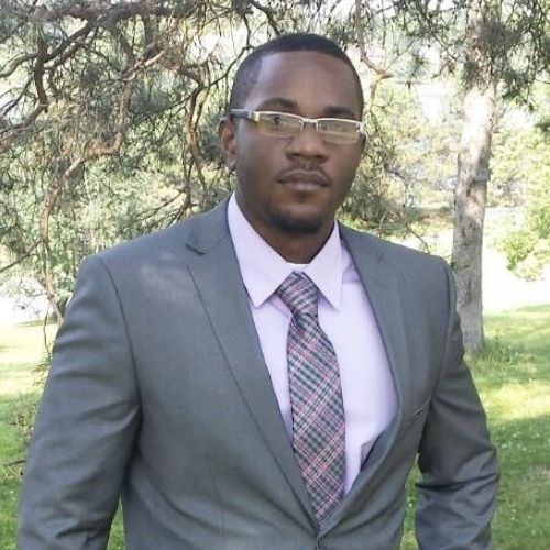 Devin has dark skin, short clipped hair, and wears rectangular glasses and a light pink shirt under his grey suit. He is standing in front of a park.