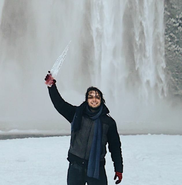 Eduardo stands in front of a frozen landscape, bundled in all black winter gear. He holds a giant icicle up in the air triumphantly.