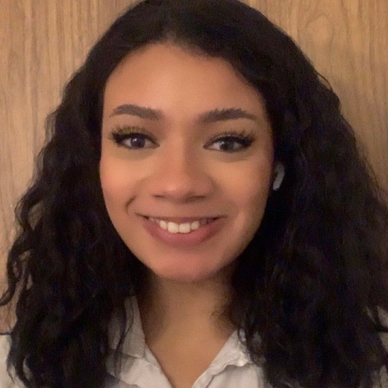 Jada, a light brown-skinned Black person with long black hair, smiles in front of a brown wood background. She is wearing a white button-up top, captured from the shoulders-up.