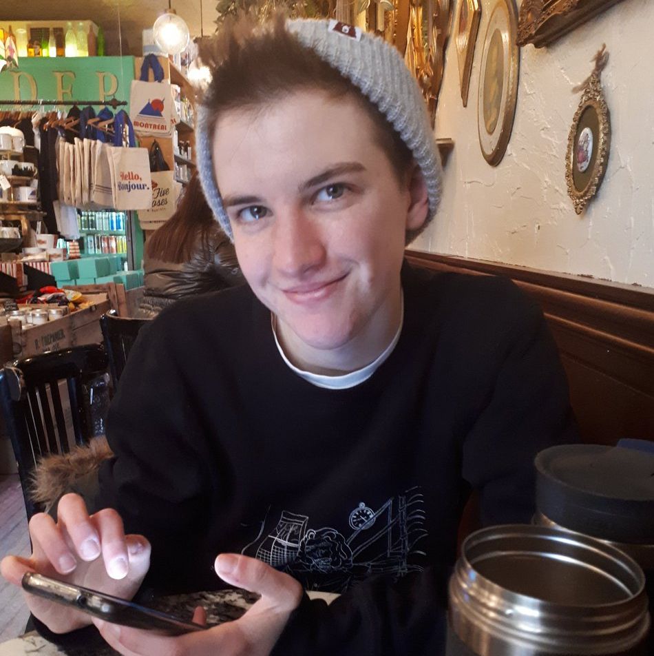 Jay, who has light skin and short brown hair under a grey touque, sits in a pub. He is looking up at the camera and smiling, his hand poised over his phone mid-tap.