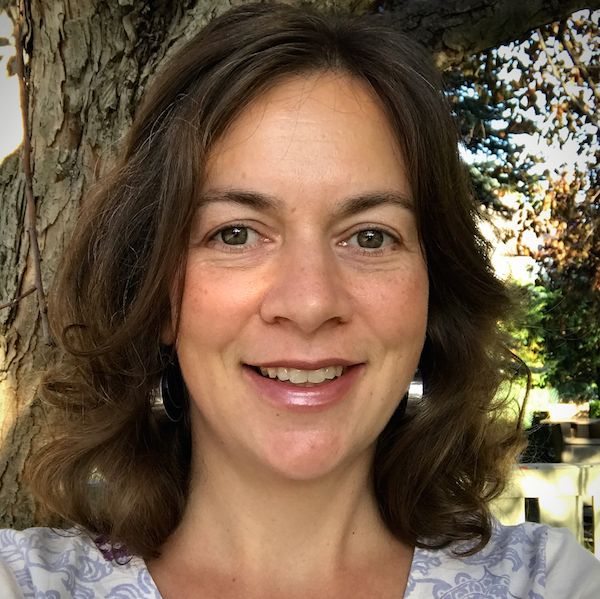 Kate stands in front of a tree and smiles at the camera. She has light skin and softly curled, shoulder-length brown hair. She is wearing silver earrings and a grey and white patterned shirt.