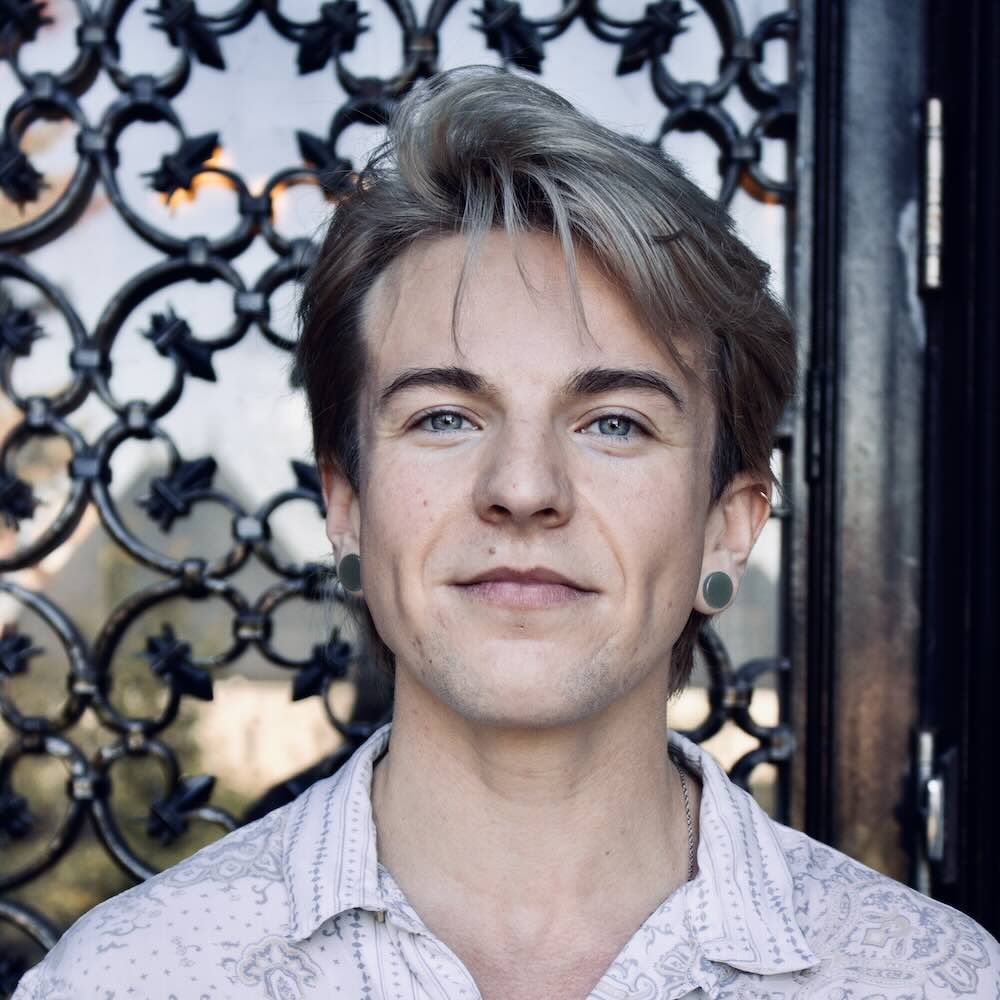 A portrait of Kit standing in front of a wrought-iron gate. He has light skin, short silver hair, a slight smile and is wearing a white button-down shirt.