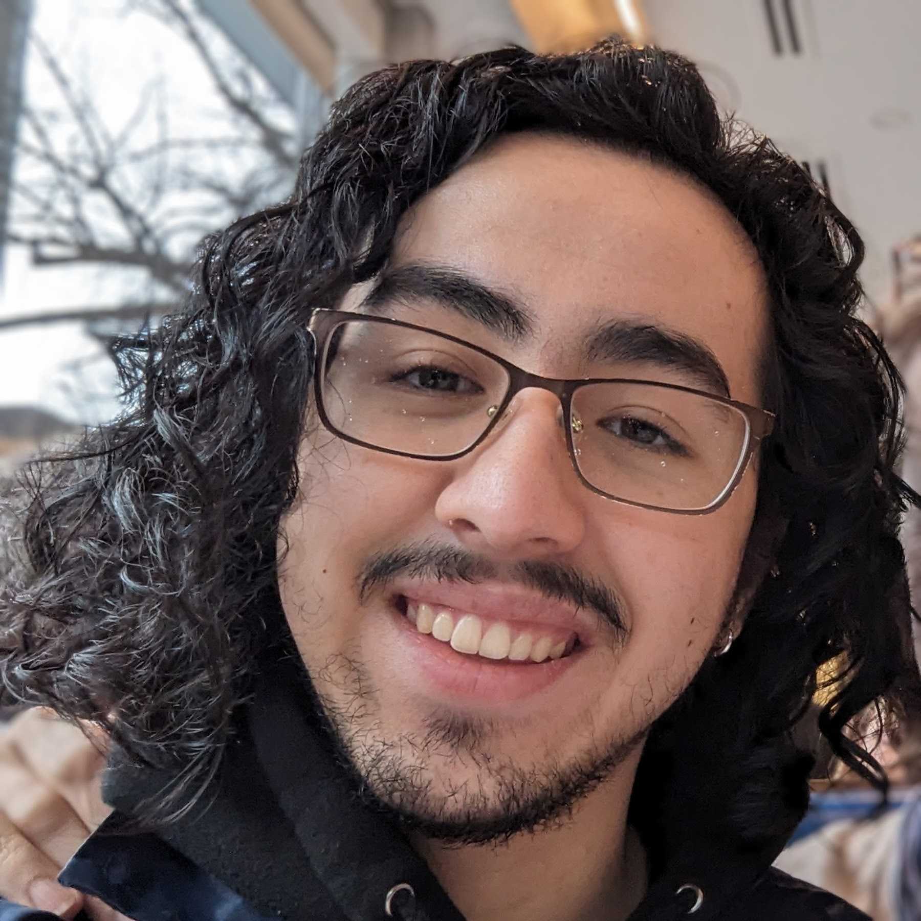 Maddie has light skin, shoulder-length curly black hair, and facial hair. He is wearing glasses and is smiling at the camera.