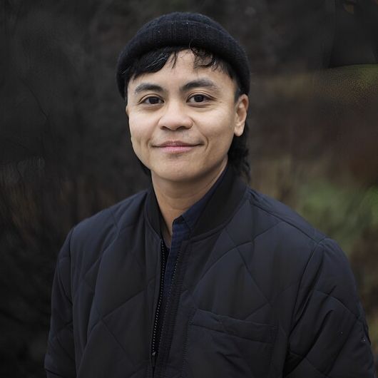 Mel has light brown skin and dimples. He wears a dark jacket, his dark hair peeking out from under his touque, standing in front of an out-of-focus forest.