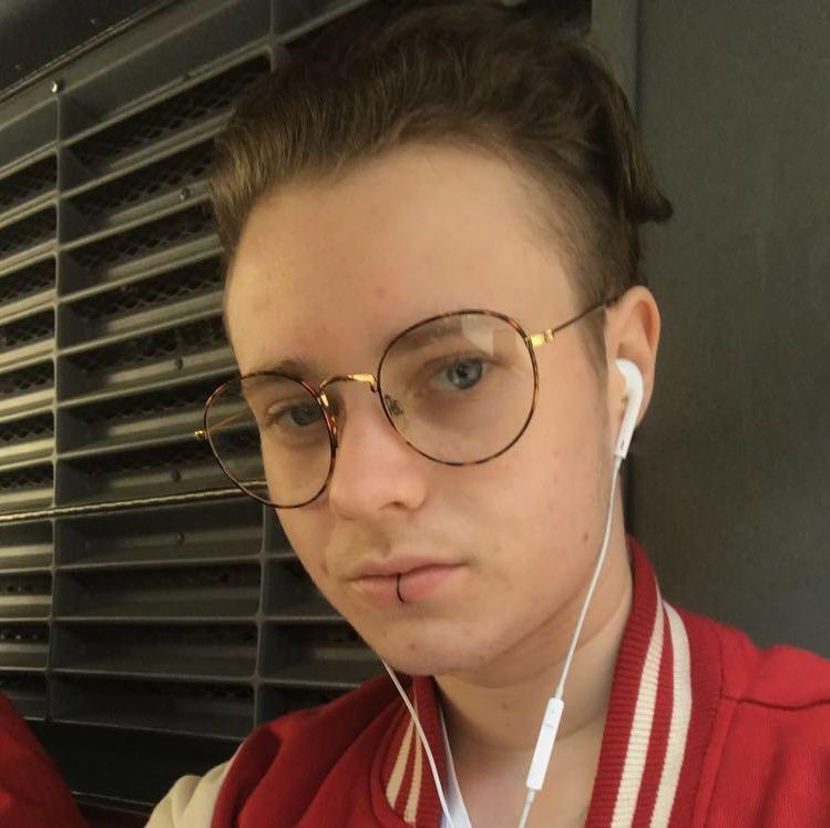 Noah has light skin and short brown hair pushed out of his face. He is wearing a red jacker, white wired headphones, large wire-rimmed glasses, and a labret lip ring.