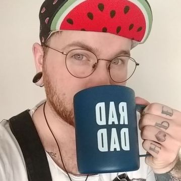 Oliver has light skin, a short, red beard and blue eyes. He is wearing a watermelon-patterned ballcap, round wire-rimmed glasses, and black overalls over a white t-shirt. He glances up at the camera, mid-sip from a mug reading RAD DAD in mirror image. The hand holding the mug as knuckle tattoos reading HOPE.