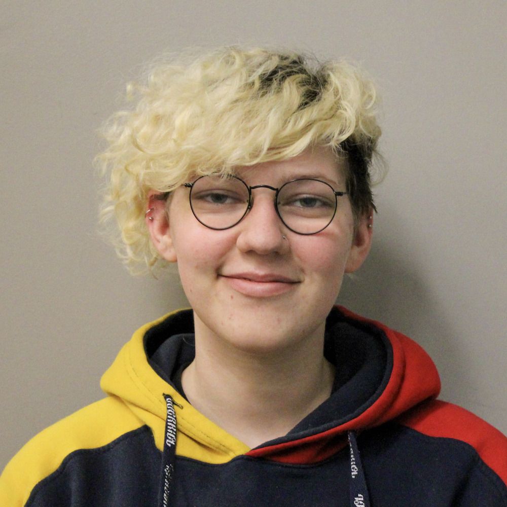 Daniel has light skin and long, bleach-blonde curly bangs. He is wearing a yellow, black, and red hoodie and glasses and is smiling at the camera.