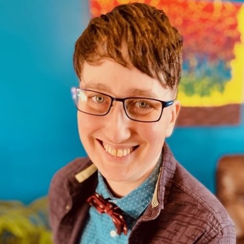 Danielle has short brown hair, light skin, and black rectangular glasses. They are wearing a brown jacket over a blue button down shirt and red polka dot bow tie, smiling at the camera in front of a colourful, out-of-focus backdrop.