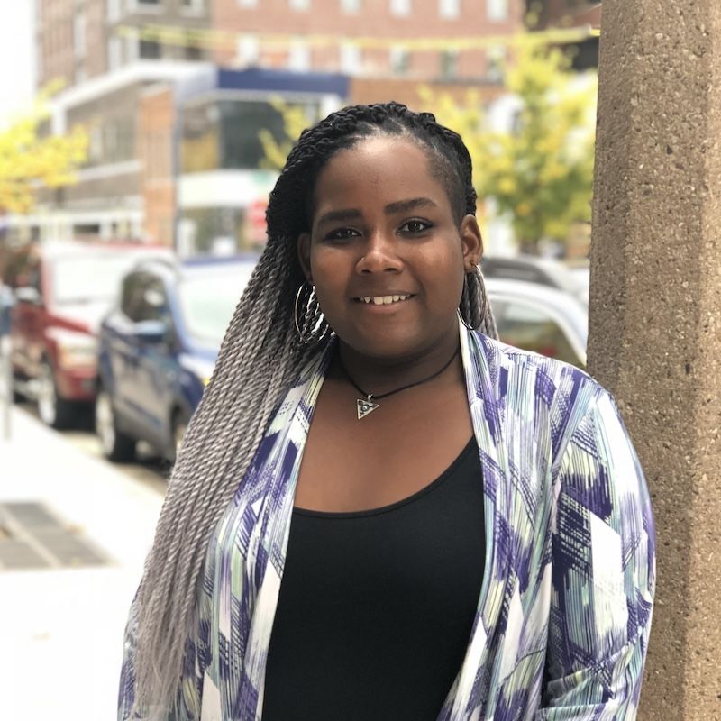 LaVelle has dark skin and long black and white braids. She is wearing a black tank top and a purple and white cardigan, smiling at the camera while standing outside on a sidewalk.