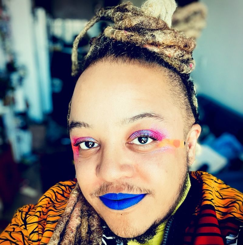 Syrus smiles in blue lipstick up at the camera. His light Black skin is complimented by vivid pink, blue, and orange makeup, and his blonde dreadlocks are tied on top of his head. He wears a colourful jacket.